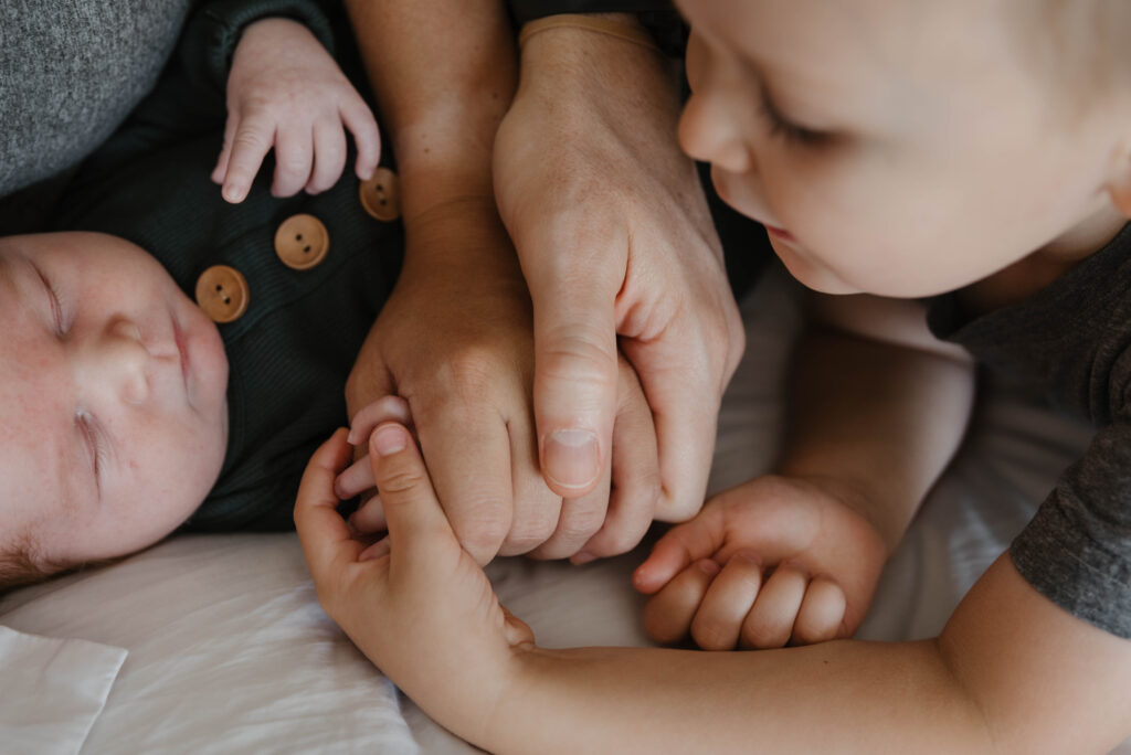 an example of baby-led posing with big brother looking over at baby and holding baby's hand on a bed with parents
